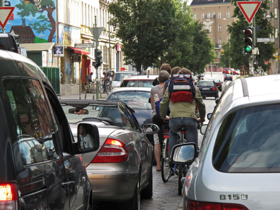 Ein Ausschnitt von stehenden Autos, durch die sich Fahrradfahrer*innen drängen. 