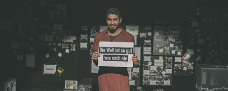 Schauspieler hält Schild "Die Welt ist so gut wie noch nie."  - Link auf: Theaterstück "Revolution: Alles wird gut!"