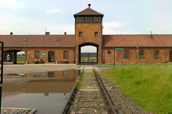 Das Bild zeigt eine Frontalaufnahme aus dem KZ Auschwitz-Birkenau. Im Hintergrund ist ein Backsteingebäude mit einer großen Einfahrt zu sehen, auf die ein langes Gleis hinführt. 